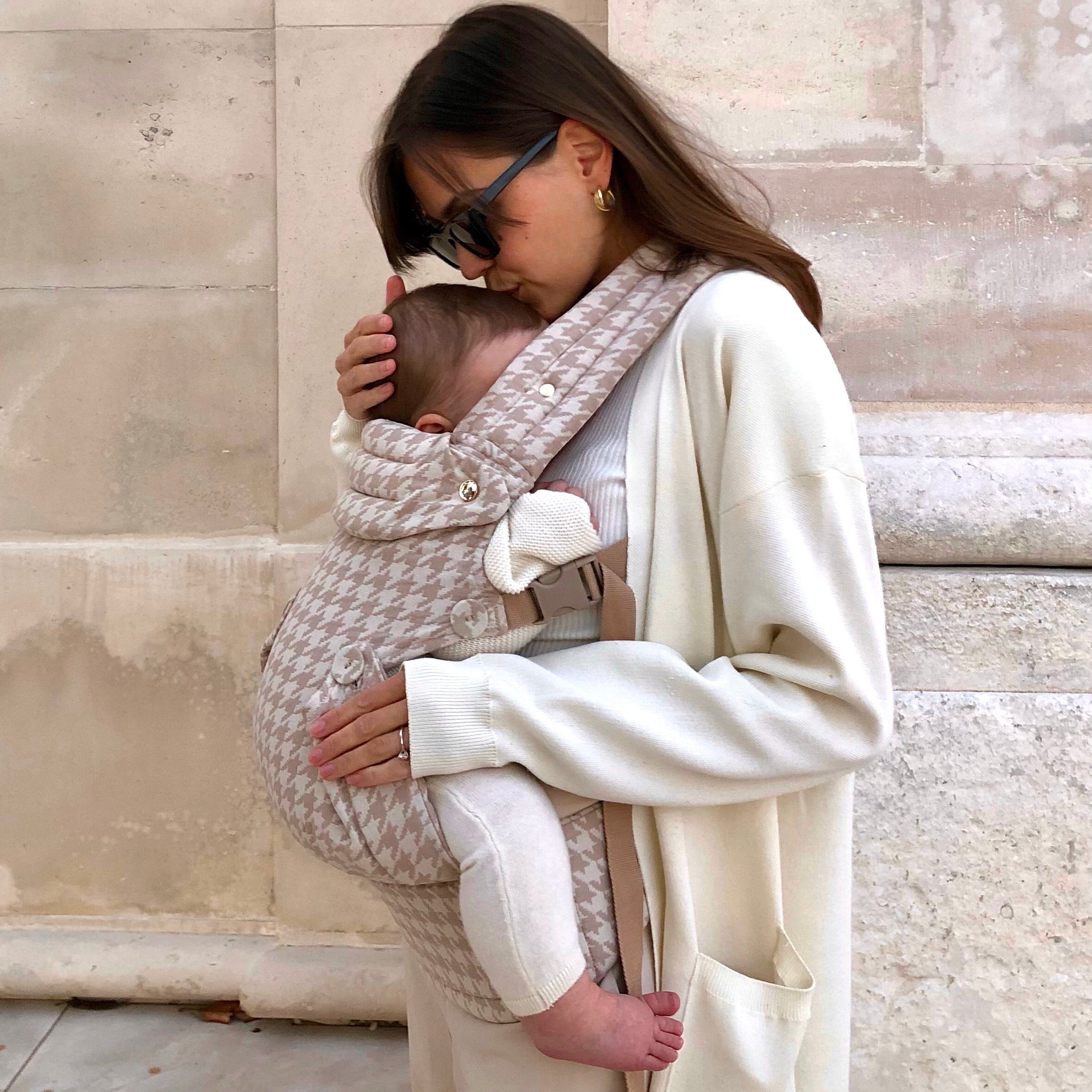 Woman holding a baby in a carrier against a stone wall.