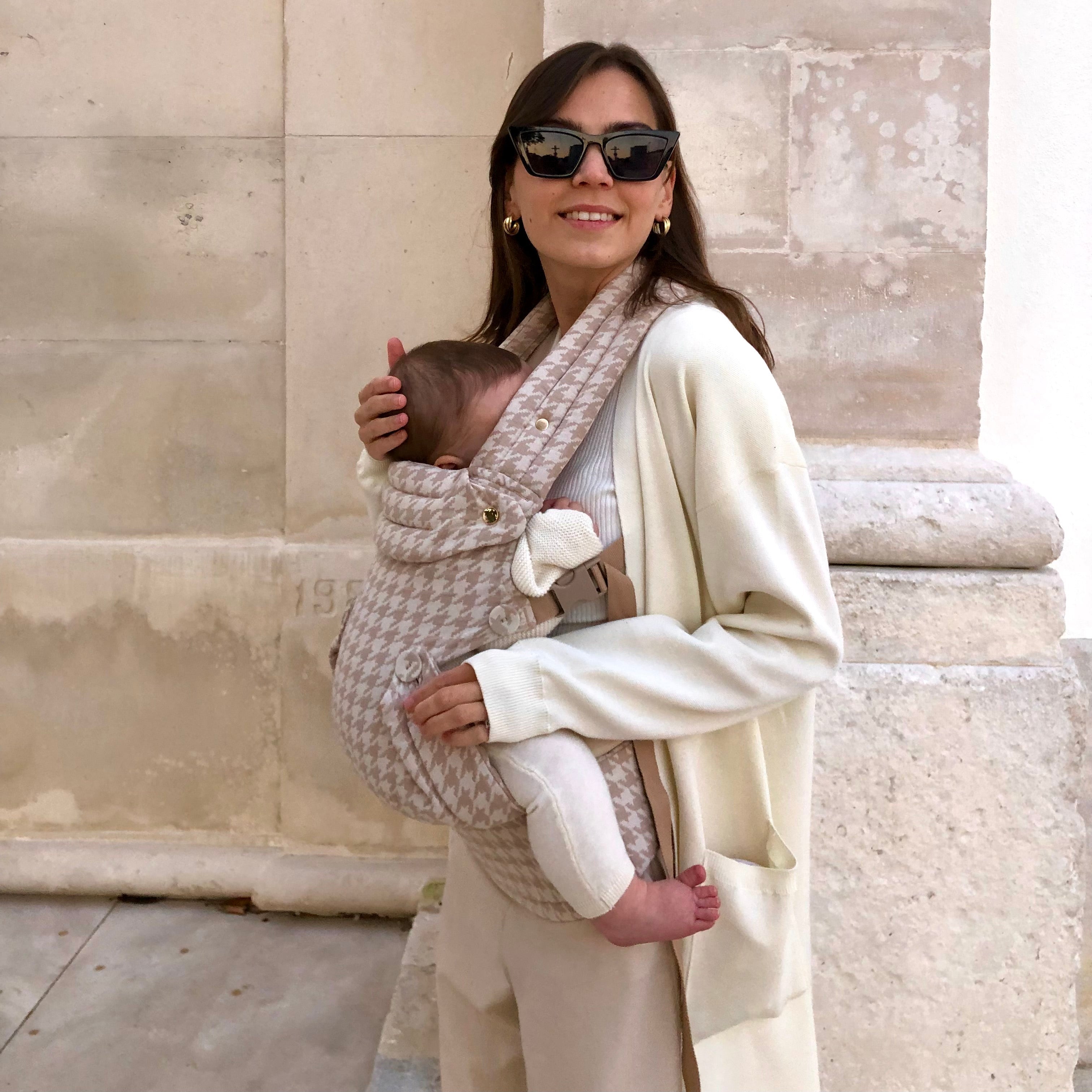 Woman holding a baby in a sling against a stone wall.